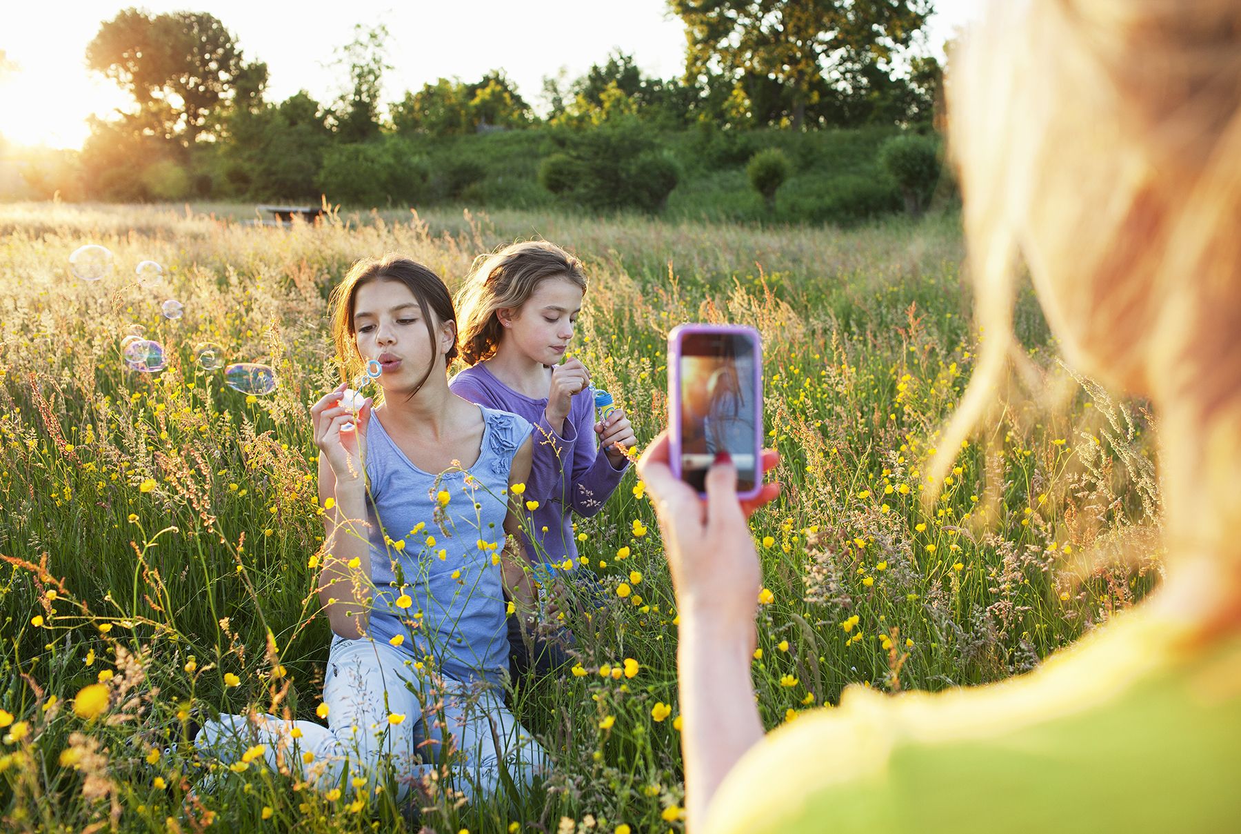 Жена снима фотографију две девојке у пољу које дувају мехуриће