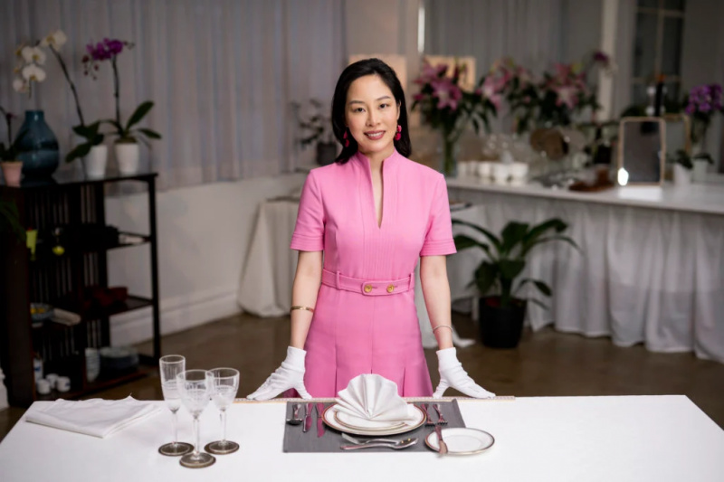   Sara Jane Ho har på seg rosa kjole og hvite hansker med hendene på middagsbordet med tallerkener og glass. Planter i bakgrunnen.